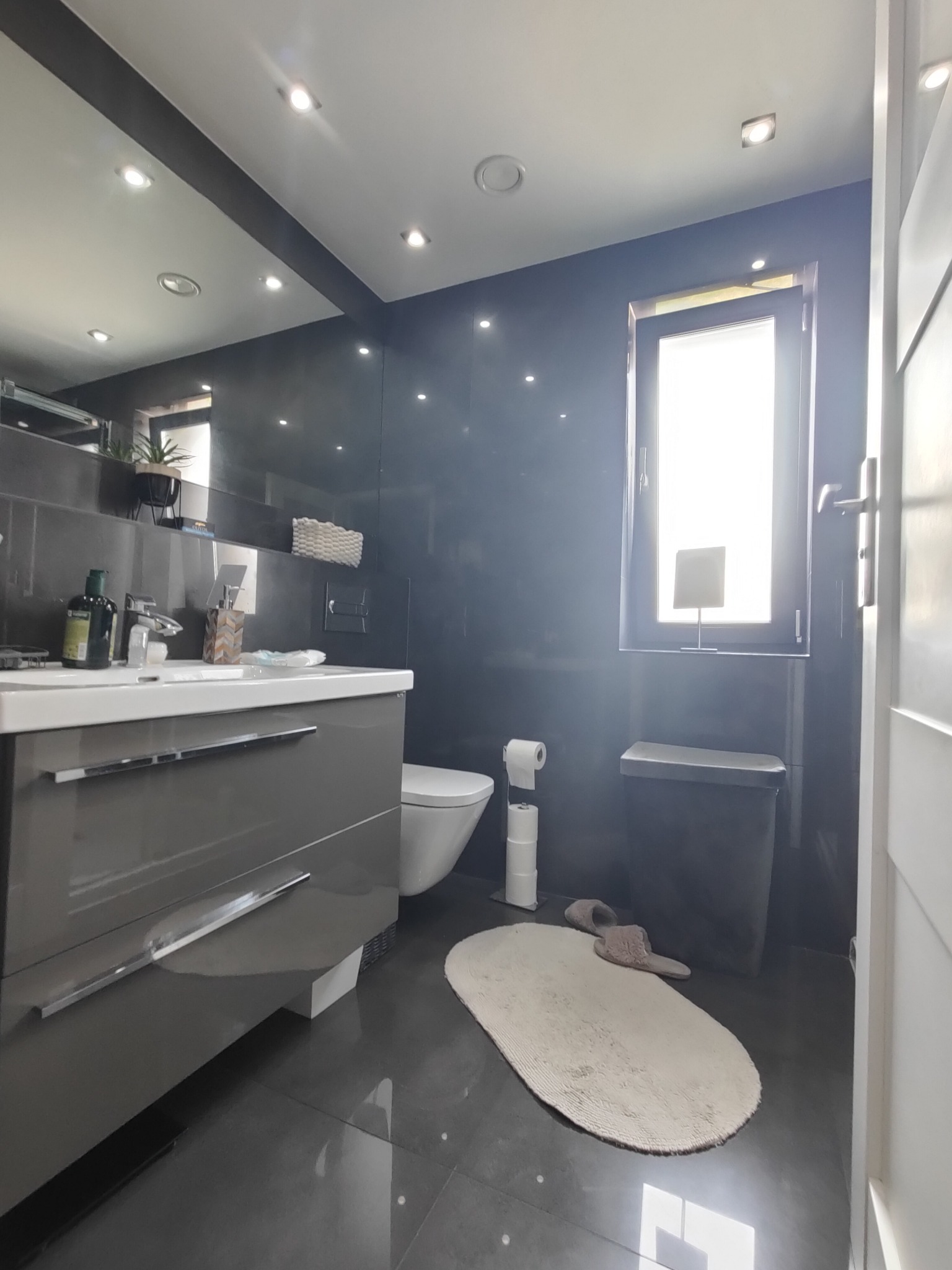 Nowoczesna łazienka z szarą armaturą, błyszczącymi płytkami podłogowymi i owalnym dywanikiem, widziana z perspektywy wejścia.