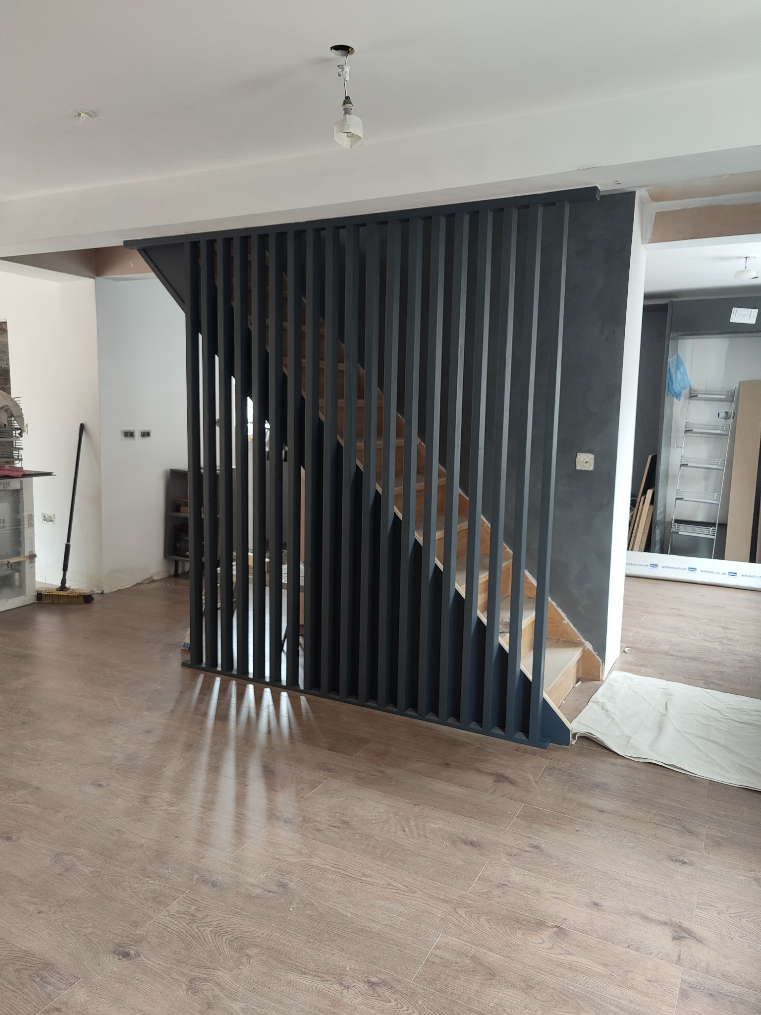 Nowoczesna drewniana klatka schodowa z ażurową, grafitową ścianką działową, widoczna na tle parkietu i surowego wykończenia wnętrza.