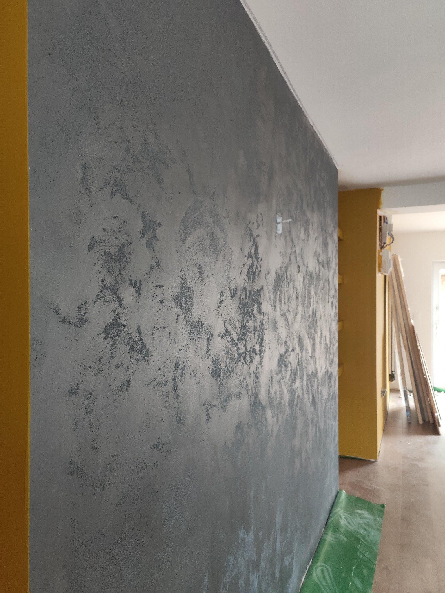 Ściana z efektem dekoracyjnym w odcieniach szarości, żółte narożniki, podłoga zabezpieczona folią ochronną, widoczne deski w tle.