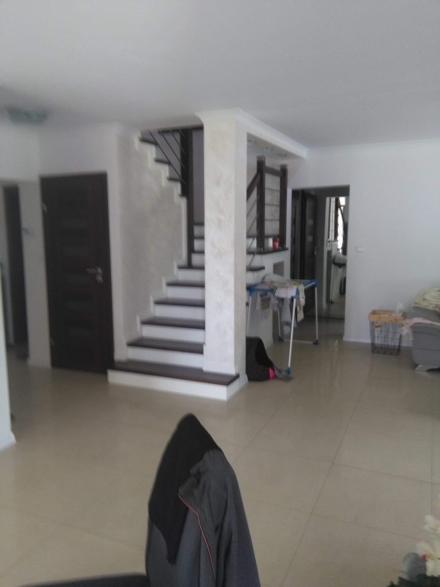 Jasne wnętrze domu z widocznymi schodami z ciemnymi stopniami, brązowymi drzwiami i beżową podłogą z płytek. W tle widoczny suszarka na pranie i inne przedmioty.