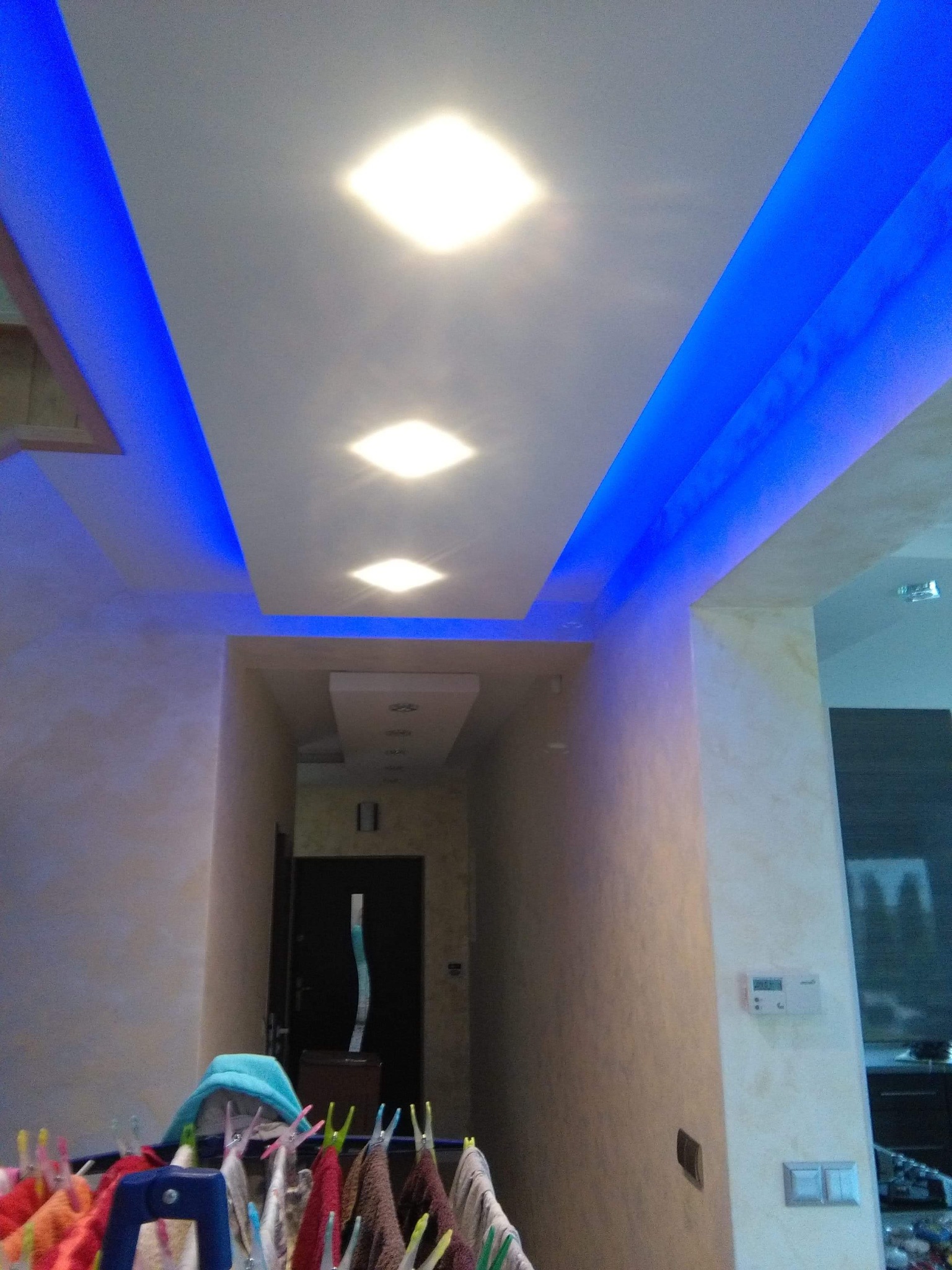 Nowoczesne oświetlenie sufitowe z niebieskimi diodami LED w korytarzu, widok z dołu na sufit podwieszany z wbudowanymi lampami kwadratowymi i dekoracyjnym oświetleniem LED, w tle widoczny suszarka...