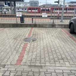 Szara kostka brukowa na parkingu z czerwoną linią wyznaczającą miejsce postojowe, w tle metalowe barierki, peron kolejowy i czerwony pociąg.