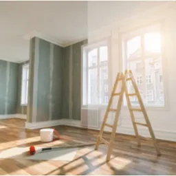 Pokój w trakcie remontu: ściany częściowo pokryte szpachlą, podłoga drewniana, drabina oparta o ścianę przy oknie, wiadro z farbą i wałek na podłodze.