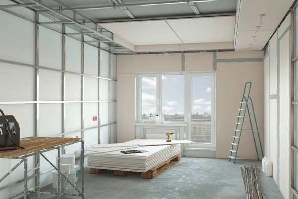 Wnętrze w trakcie montażu ścian działowych z płyt gipsowo-kartonowych na metalowej konstrukcji; widoczne płyty na palecie, drabina i narzędzia.