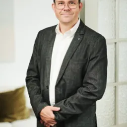 Portret mężczyzny w okularach, ubrany w szarą marynarkę i białą koszulę, stojącego w jasnym wnętrzu obok szklanych drzwi.