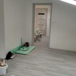 Pokój na poddaszu w trakcie remontu: świeżo pomalowane ściany na biało, panele podłogowe imitujące drewno, drzwi wewnętrzne z szarą ościeżnicą, narzędzia i materiały budowlane porozrzucane po...