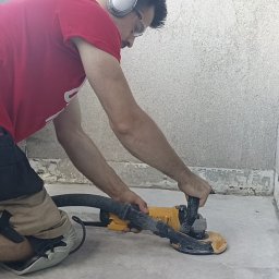 Konsolvo - Perfekcyjne wnętrza - Pracownik w słuchawkach szlifuje betonową podłogę przy użyciu szlifierki z odkurzaczem, widoczny pas z narzędziami i nóż.