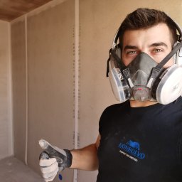 Konsolvo - Perfekcyjne wnętrza - Pracownik w masce ochronnej i słuchawkach, ubrudzony pyłem, kciukiem w górę na tle surowych ścian z płyt gipsowo-kartonowych przed szpachlowaniem, widoczne logo firmy na koszulce.
