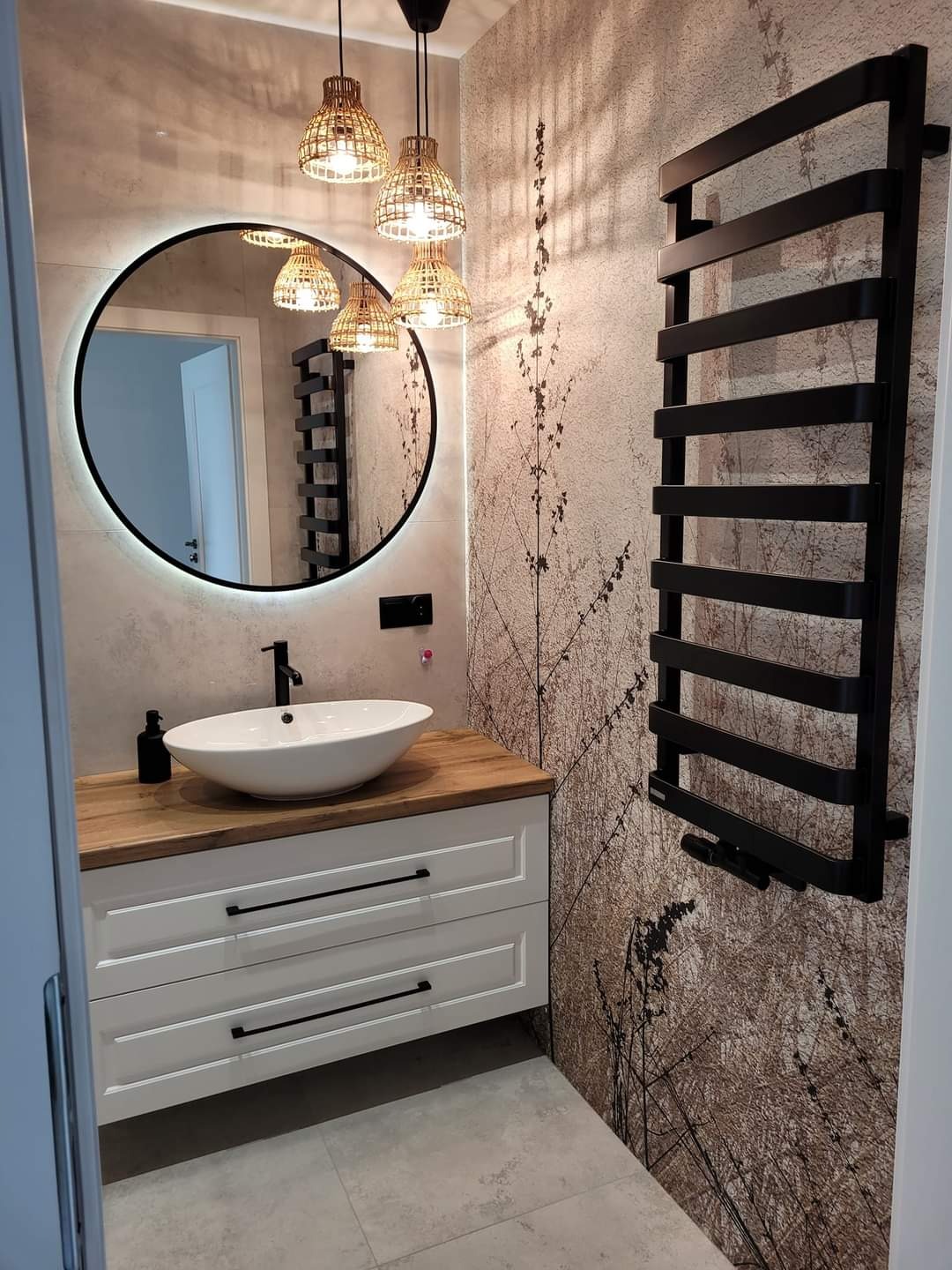 Nowoczesna łazienka z owalną umywalką nablatową na drewnianym blacie, białą szafką z czarnymi uchwytami, okrągłym lustrem z podświetleniem LED, czarnym grzejnikiem drabinkowym i tapetą z motywem...