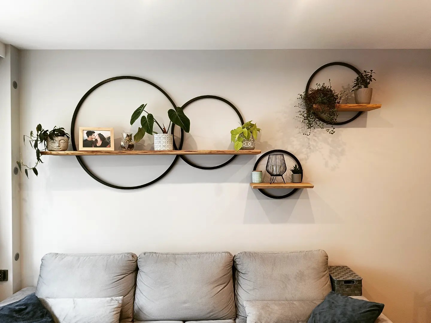 Ściana salonu z trzema okrągłymi półkami z drewnianymi blatami, na których ustawione są rośliny doniczkowe, ramka ze zdjęciem i dekoracje, nad szarą sofą.