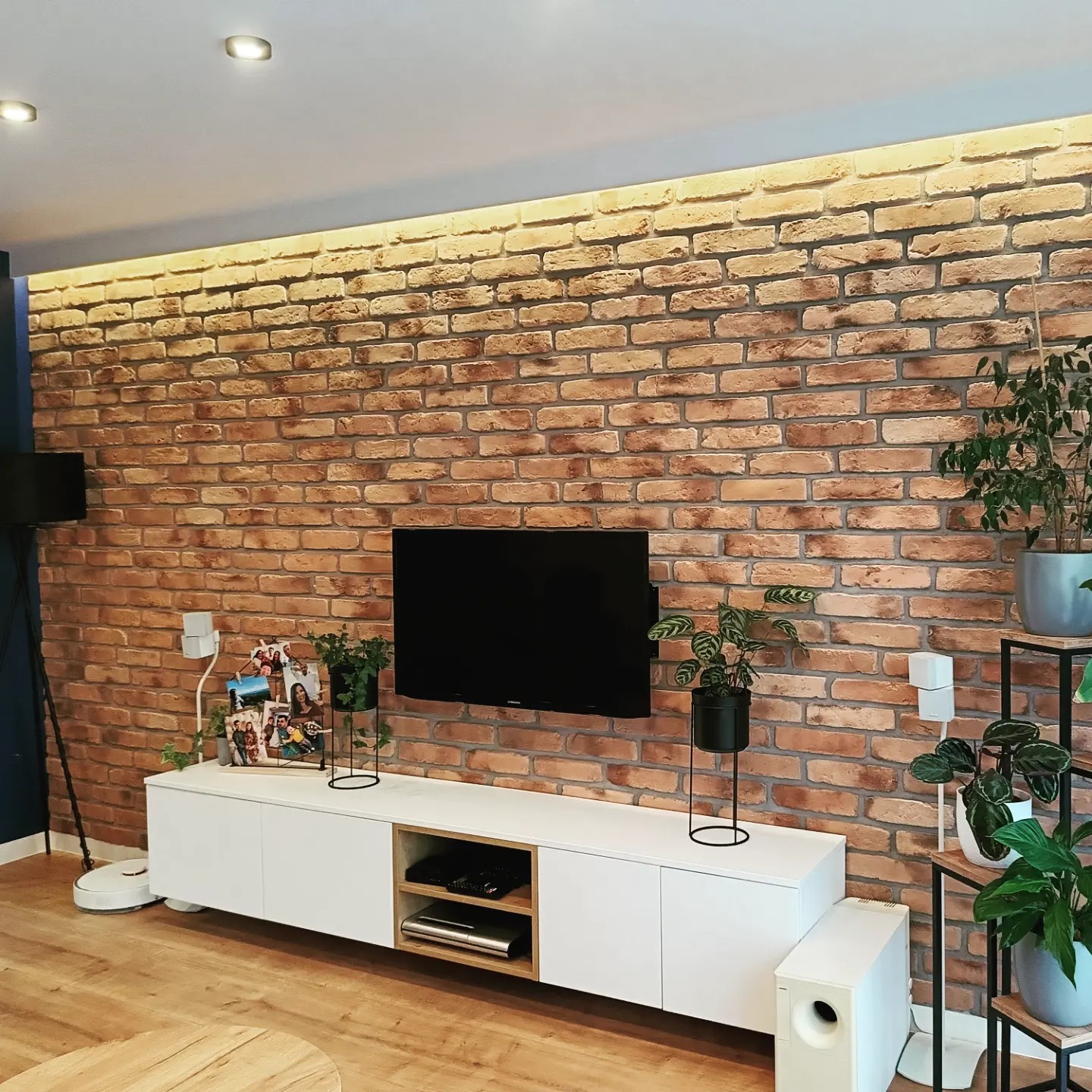 Salon z ceglaną ścianą podświetloną od góry, biała szafka RTV z dekoracjami, roślinami i telewizorem, drewniana podłoga.