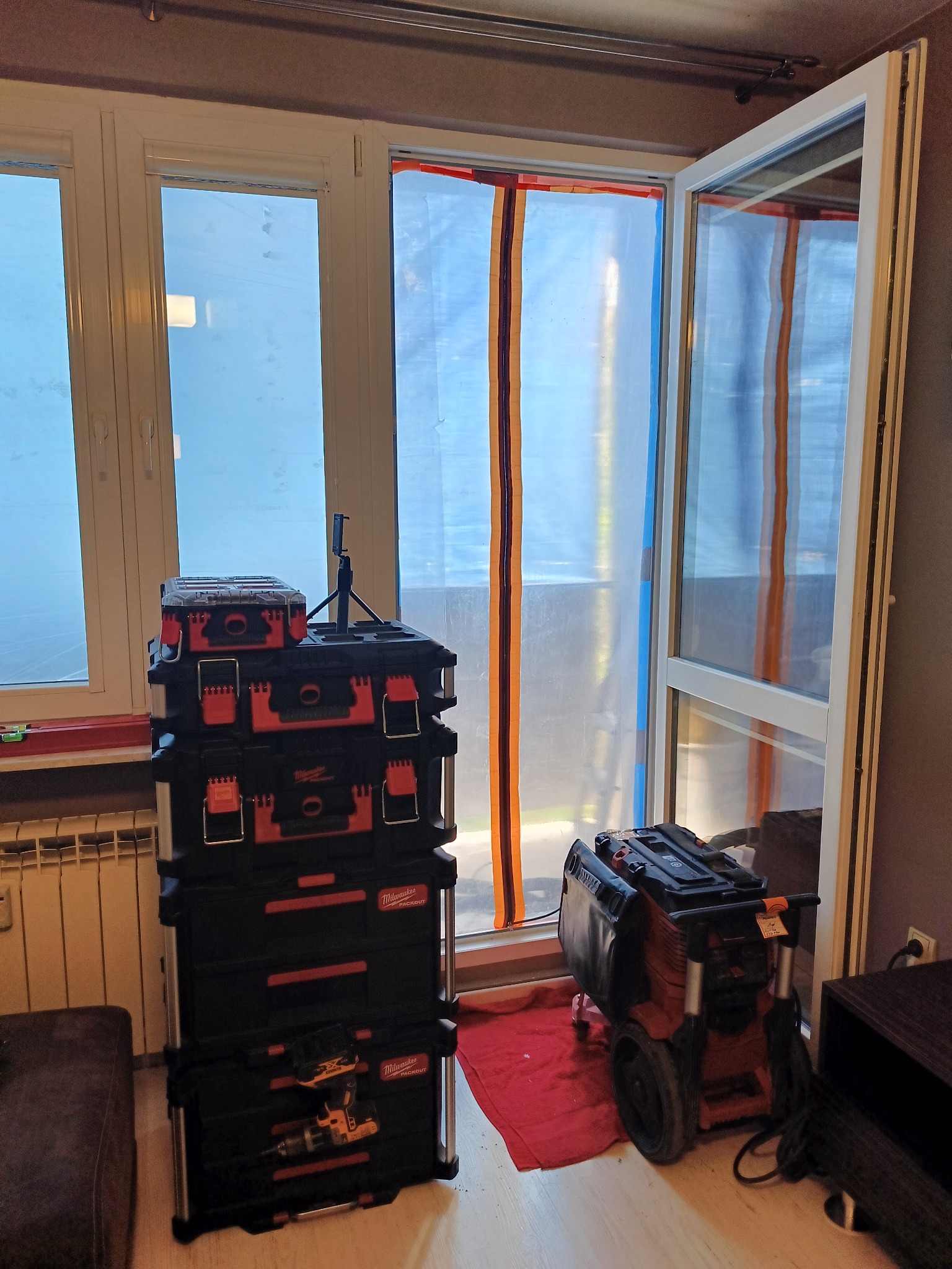 Przygotowania do remontu: okno zabezpieczone folią malarską z taśmą, obok ustawione skrzynki narzędziowe Milwaukee Packout i odkurzacz przemysłowy na kółkach