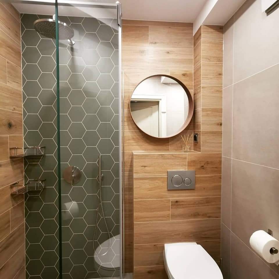 Nowoczesna łazienka z kabiną prysznicową wyłożoną heksagonalnymi płytkami w odcieniach szarości, drewnopodobnymi płytkami na ścianach, okrągłym lustrem w drewnianej ramie i podwieszaną toaletą...