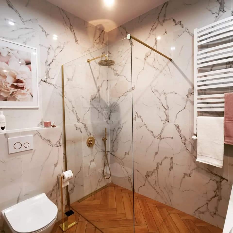 Nowoczesna łazienka z kabiną prysznicową typu walk-in, wykończona białymi płytkami imitującymi marmur, złotą armaturą i drewnianą podłogą ułożoną w jodełkę.