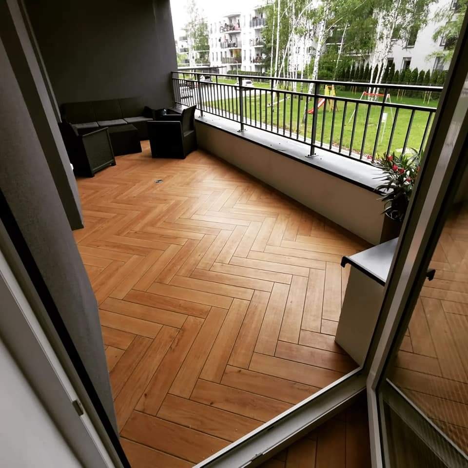 Balkon z drewnopodobną podłogą ułożoną w jodełkę, czarne meble ogrodowe i widok na plac zabaw w tle.