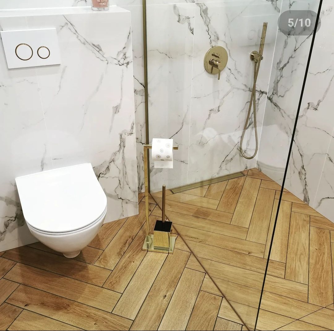 Nowoczesna łazienka z białymi płytkami imitującymi marmur, drewnopodobną podłogą ułożoną w jodełkę, złotą armaturą i akcesoriami oraz wiszącą miską WC z podtynkową spłuczką.