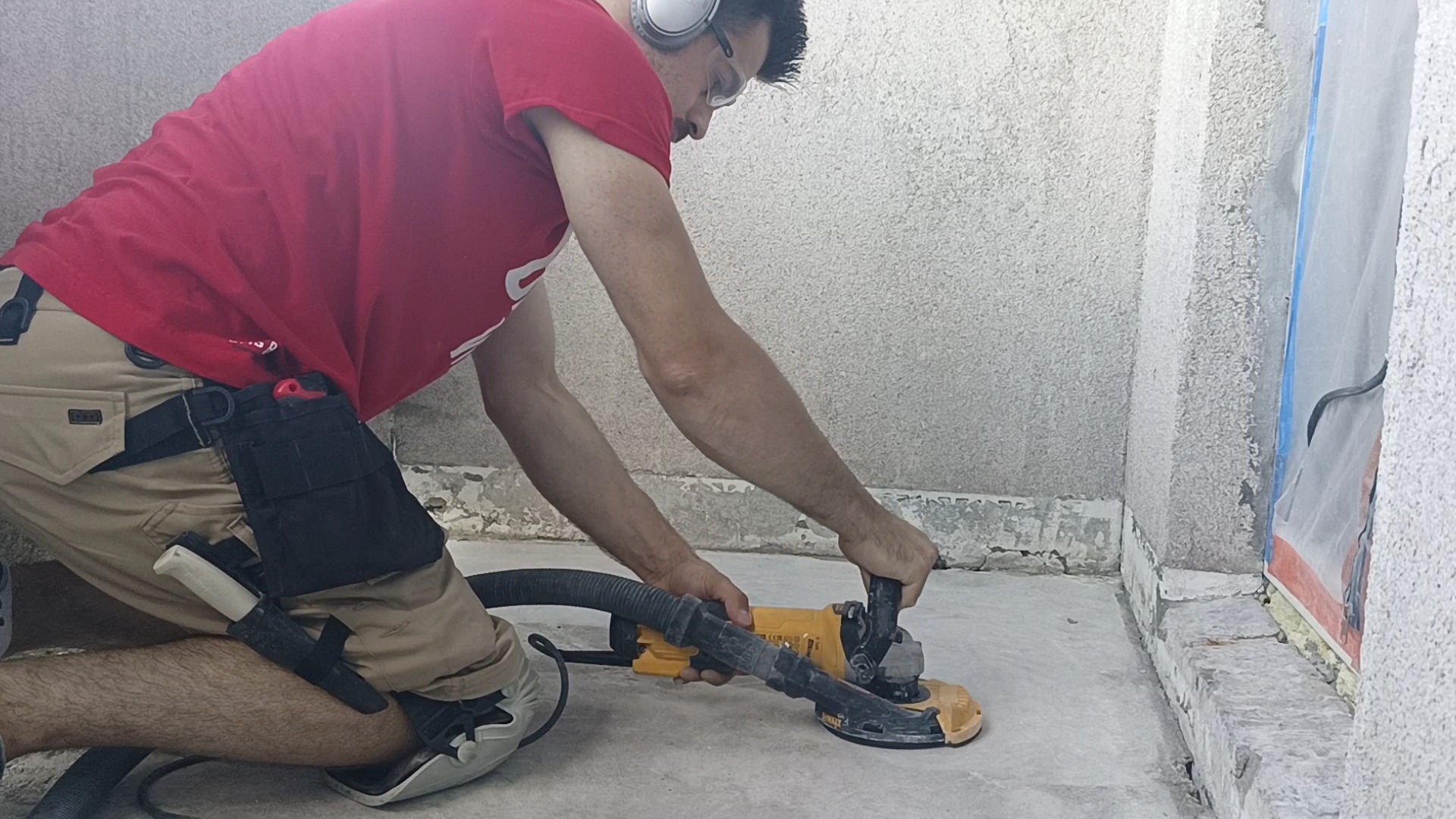 Pracownik w słuchawkach szlifuje betonową podłogę przy użyciu szlifierki z odkurzaczem, widoczny pas z narzędziami i nóż.