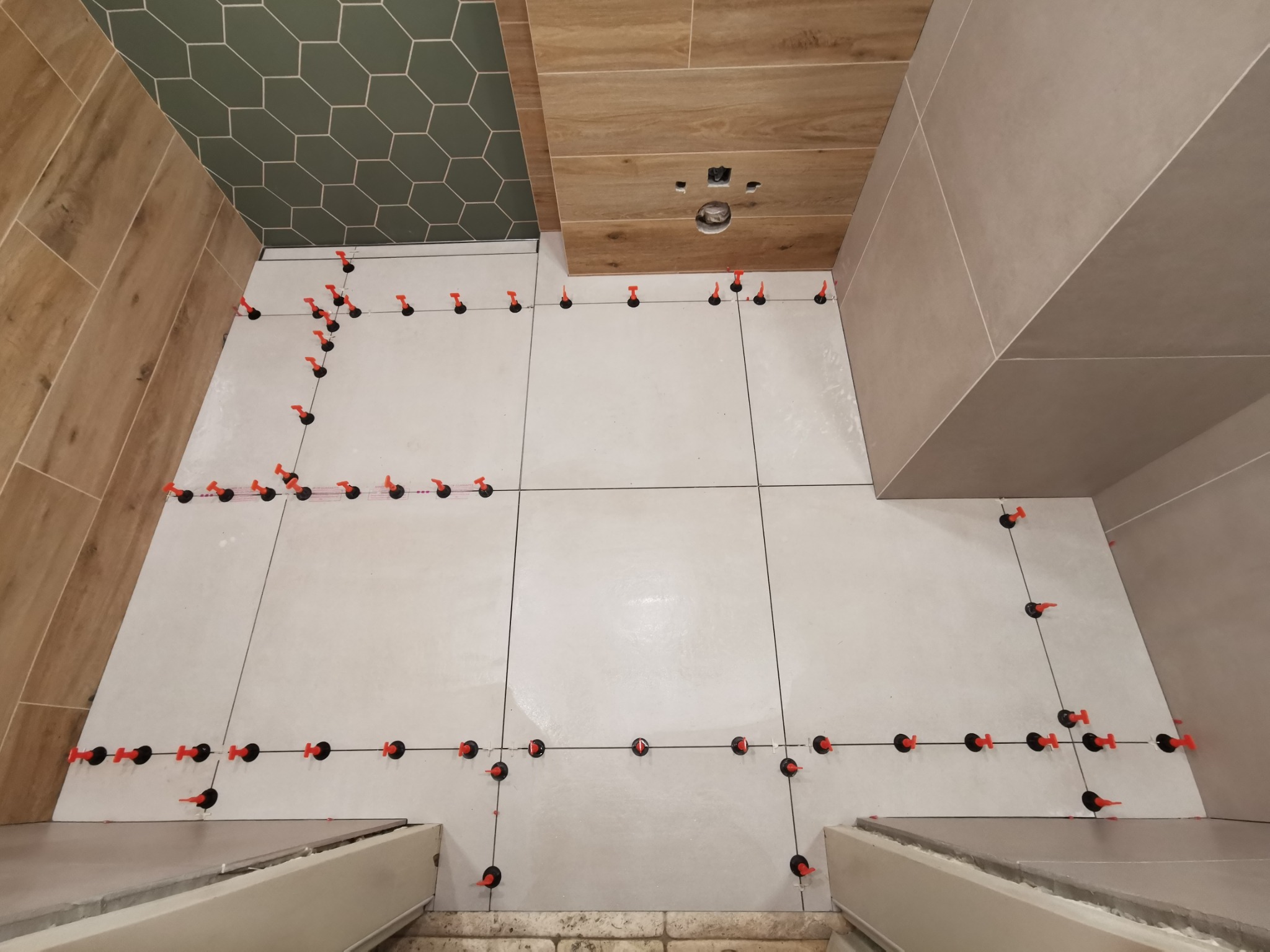 Ułożone jasne płytki podłogowe z systemem poziomowania płytek, widoczne płytki drewnopodobne na ścianie i heksagonalne płytki w odcieniach zieleni na górze ściany.