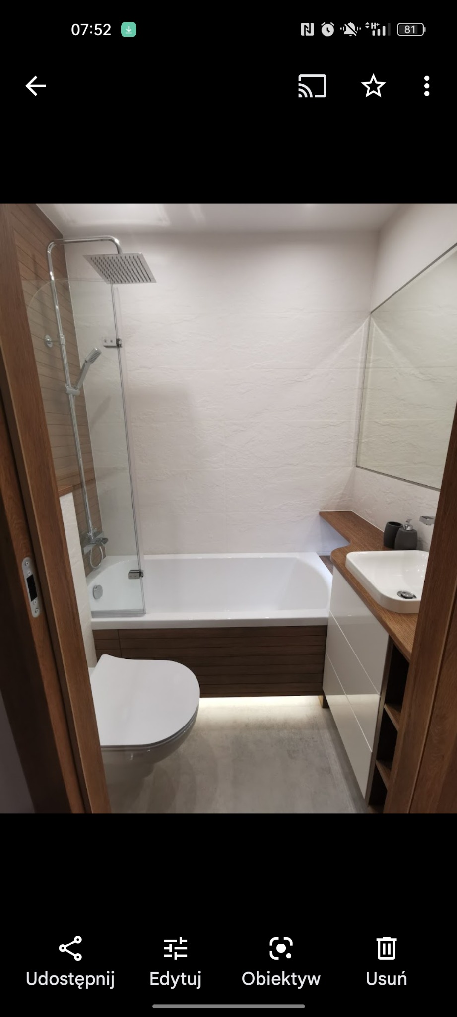 Nowoczesna łazienka z podświetlaną wanną, białą armaturą i drewnianymi akcentami, widoczna przez otwarte drewniane drzwi.