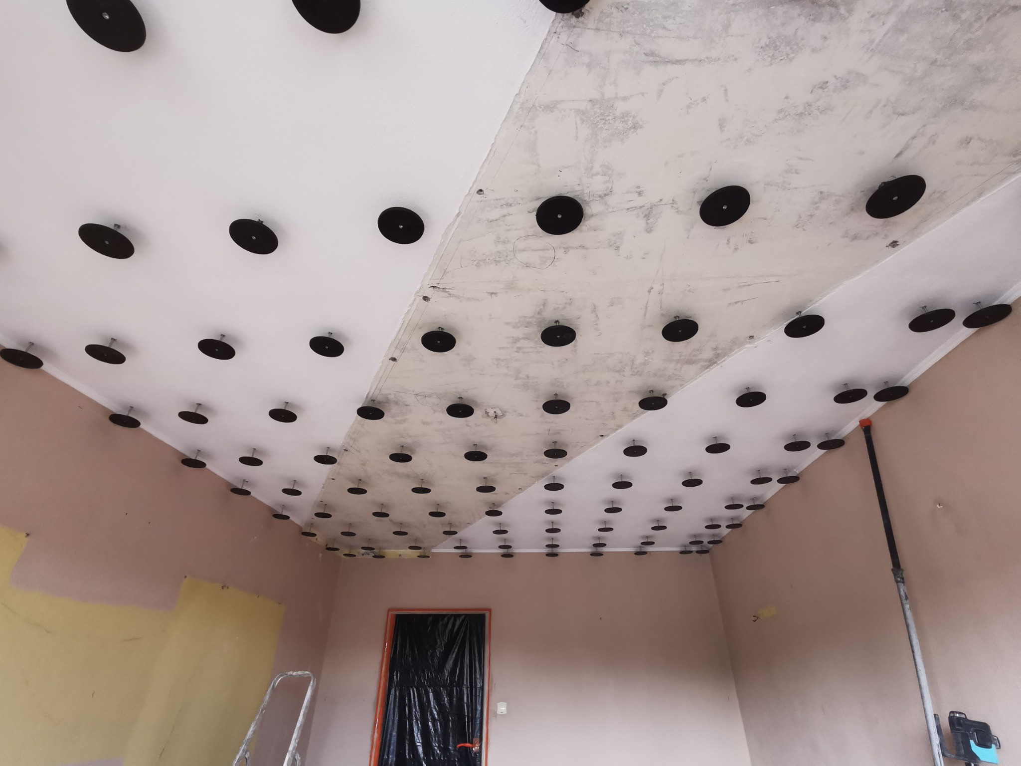 Przygotowanie sufitu do szpachlowania: widoczne czarne dyski mocujące siatkę na suficie, częściowo pokrytym szpachlą, zarys drzwi zabezpieczonych folią, drabina w rogu pomieszczenia.