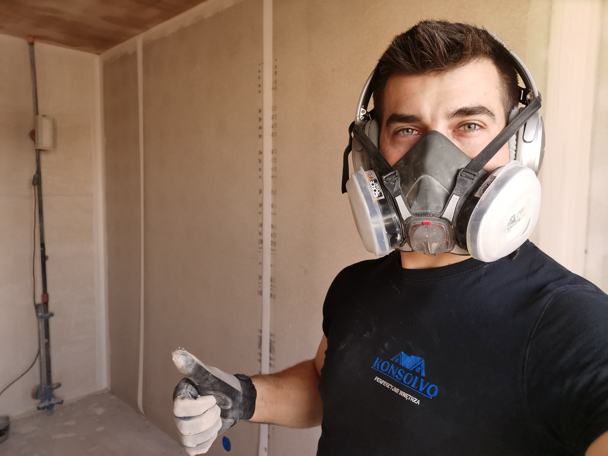 Pracownik w masce ochronnej i słuchawkach, ubrudzony pyłem, kciukiem w górę na tle surowych ścian z płyt gipsowo-kartonowych przed szpachlowaniem, widoczne logo firmy na koszulce.