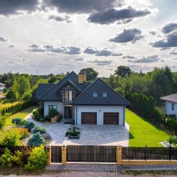 HS House Safe nieruchomości sp. z o.o. - Dwupiętrowy dom z ciemnoszarą dachówką, jasną elewacją, dwoma garażami i zadbanym ogrodem, widok z góry w słoneczny dzień z chmurami.