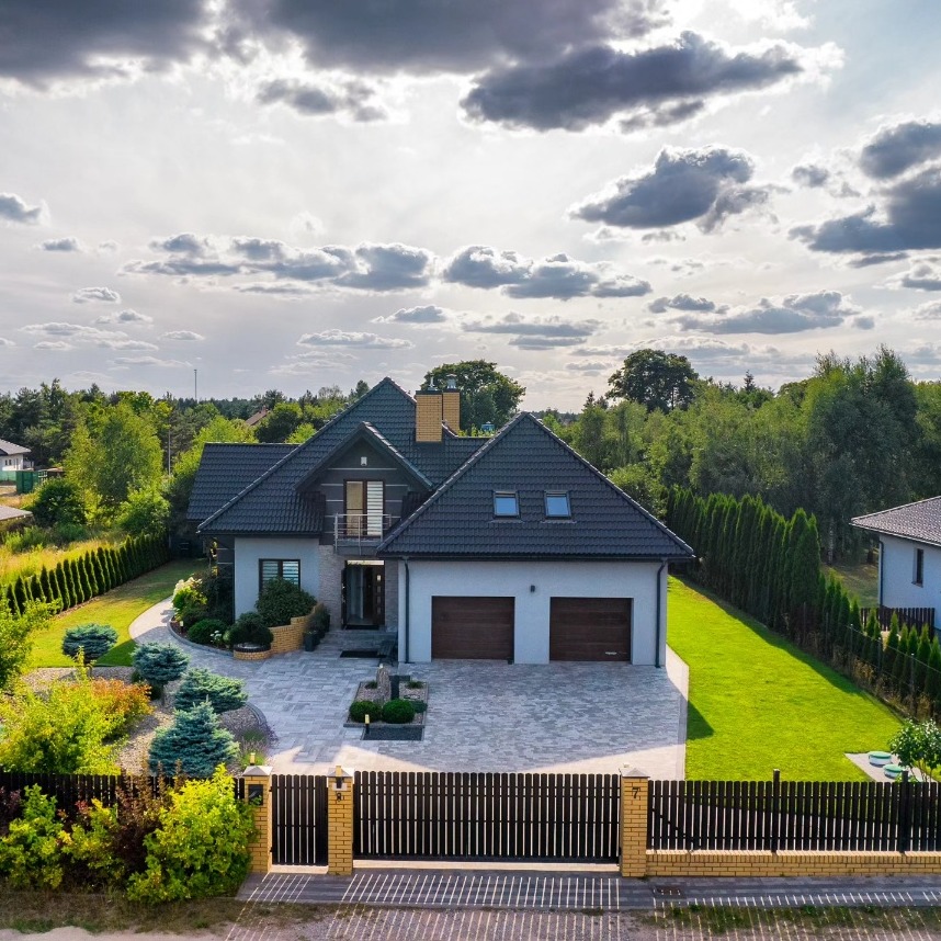 Dwupiętrowy dom z ciemnoszarą dachówką, jasną elewacją, dwoma garażami i zadbanym ogrodem, widok z góry w słoneczny dzień z chmurami.