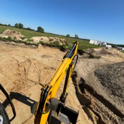 Żółta koparka JCB z widocznym ramieniem i łyżką, wykonująca wykop w piaszczystym gruncie, w tle zielone pole i budynek w trakcie budowy w słoneczny dzień.