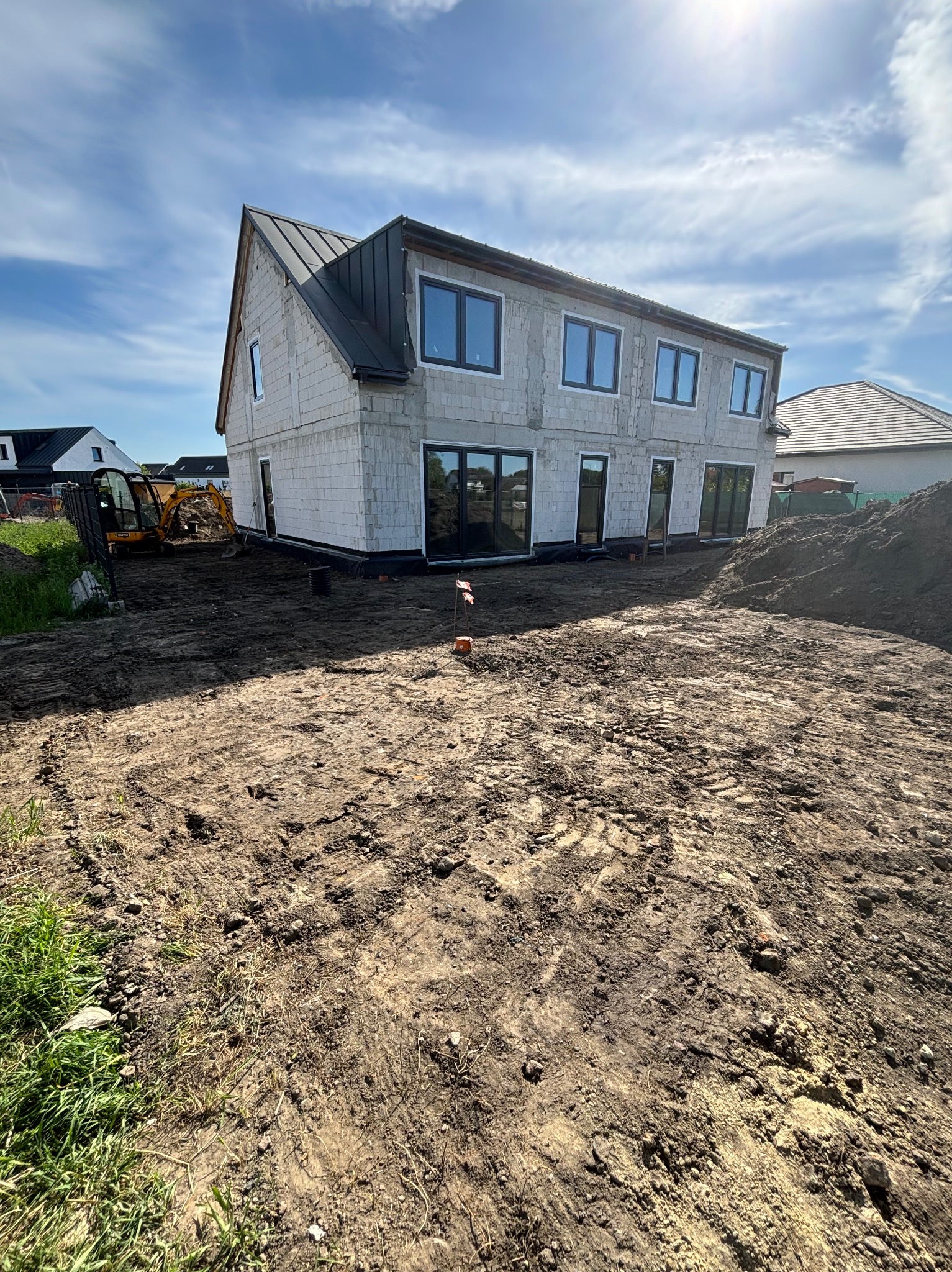 Surowy stan działki z widocznym budynkiem w trakcie budowy, teren wyrównany, w tle mała koparka.
