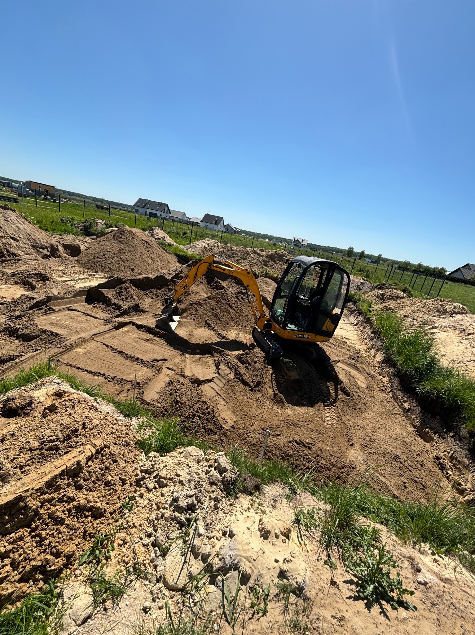 Żółta minikoparka w trakcie prac ziemnych, wyrównywania terenu pod fundamenty, widoczne ślady gąsienic i pryzmy ziemi, w tle zabudowa jednorodzinna.
