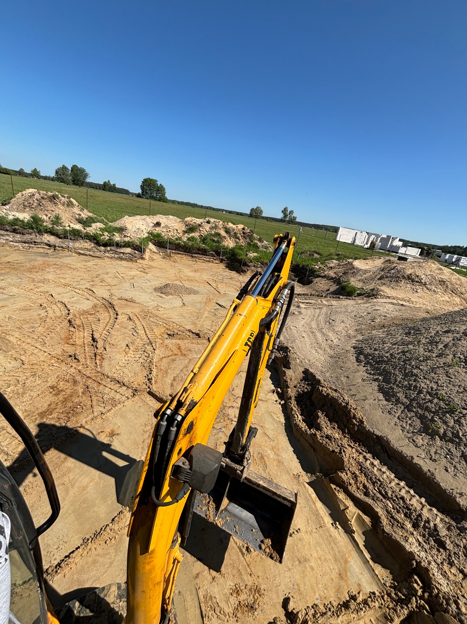 Żółta koparka JCB z widocznym ramieniem i łyżką, wykonująca wykop w piaszczystym gruncie, w tle zielone pole i budynek w trakcie budowy w słoneczny dzień.