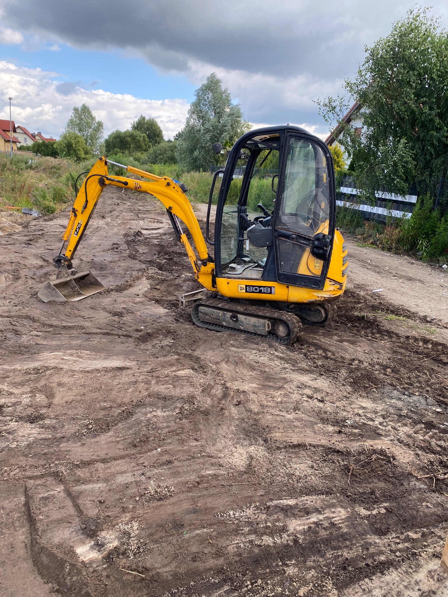 Żółta mini koparka JCB 8018 na gąsienicach, stojąca na ubitej, błotnistej ziemi, przygotowanej pod budowę, z widocznymi śladami opon i łyżką skierowaną w dół. W tle domy i drzewa.