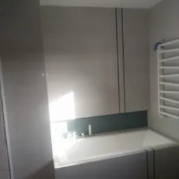 Nowoczesna łazienka w szarościach z białą wanną obudowaną panelami z czarnymi akcentami, grzejnik drabinkowy i punktowe oświetlenie sufitowe.