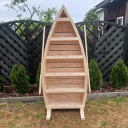 Drewniany regał w kształcie łodzi z półkami, stojący na trawniku przed czarnym ażurowym płotem i iglakami.