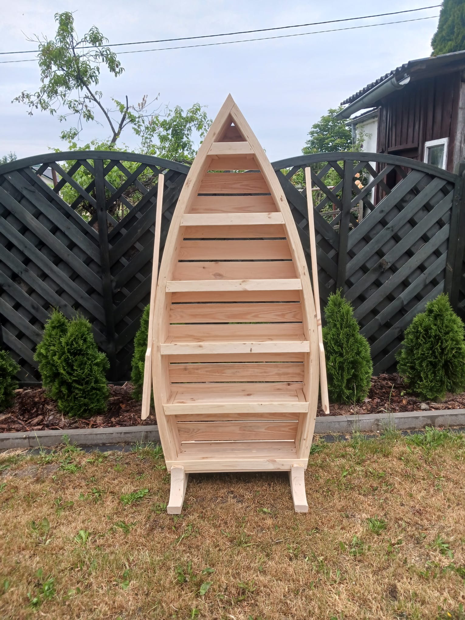 Drewniany regał w kształcie łodzi z półkami, stojący na trawniku przed czarnym ażurowym płotem i iglakami.