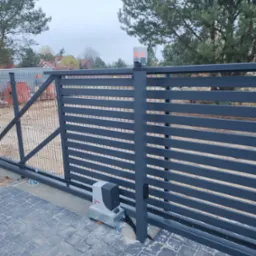 Nowoczesna, przesuwna brama w kolorze grafitowym z automatyką, zamontowana na posesji z kostką brukową i siatką ogrodzeniową w tle.