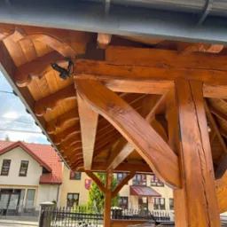 Drewniana konstrukcja altany z widocznymi elementami wykończeniowymi, zabezpieczona czujnikiem ruchu i rynną w tle budynki mieszkalne.