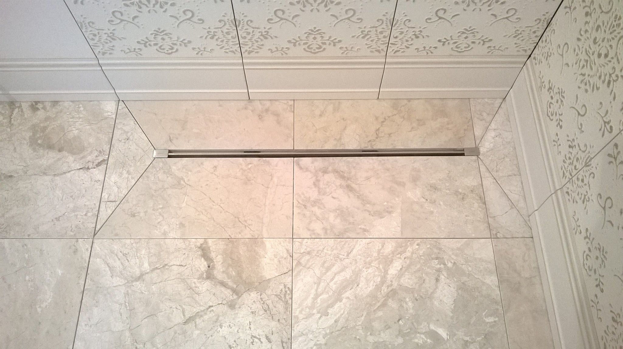Beżowe marmurowe płytki podłogowe i ścienne w łazience z liniowym odpływem prysznicowym, dekoracyjny wzór na ścianie i suficie.