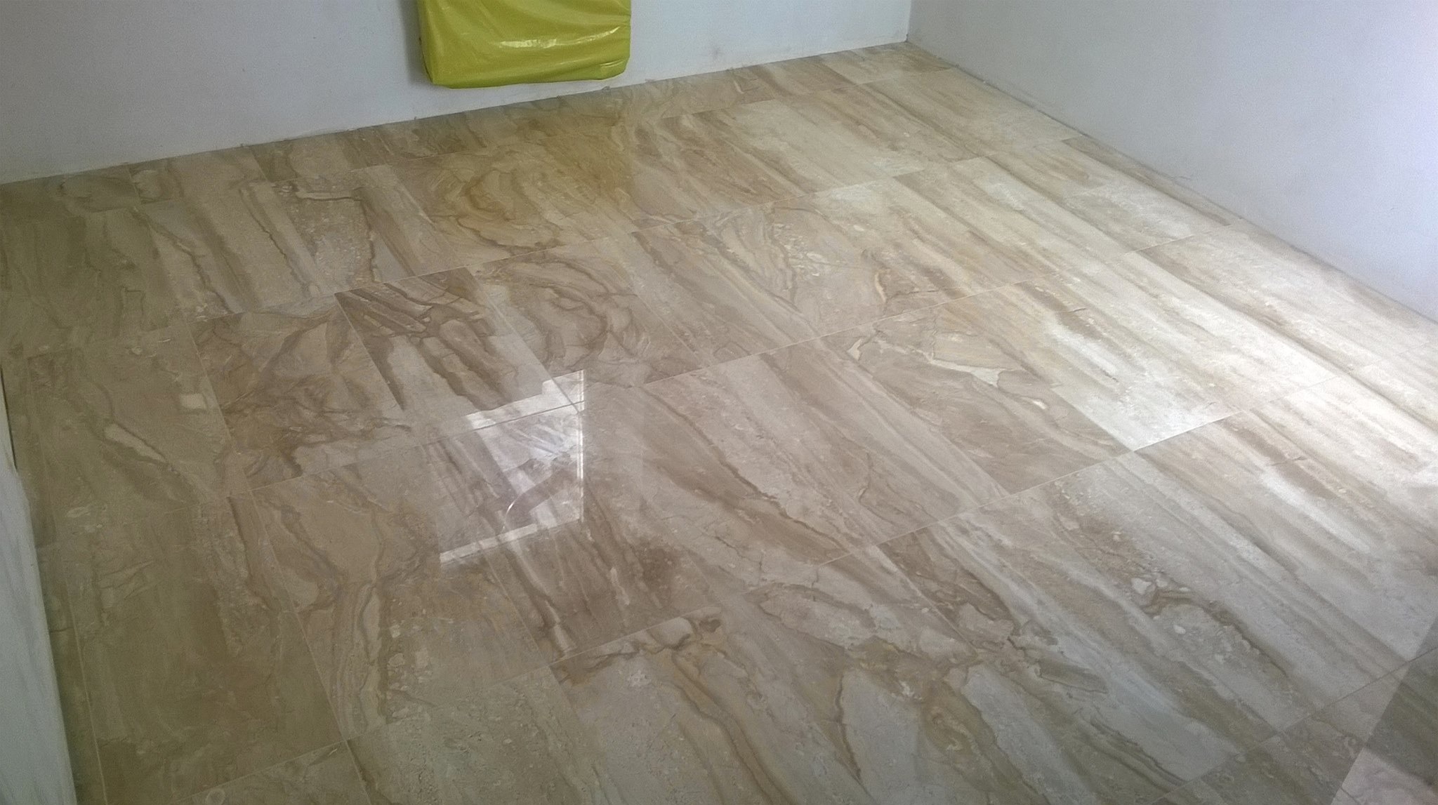 Ułożona, beżowa podłoga z płytek imitujących marmur, widoczny fragment ściany i żółta folia zabezpieczająca