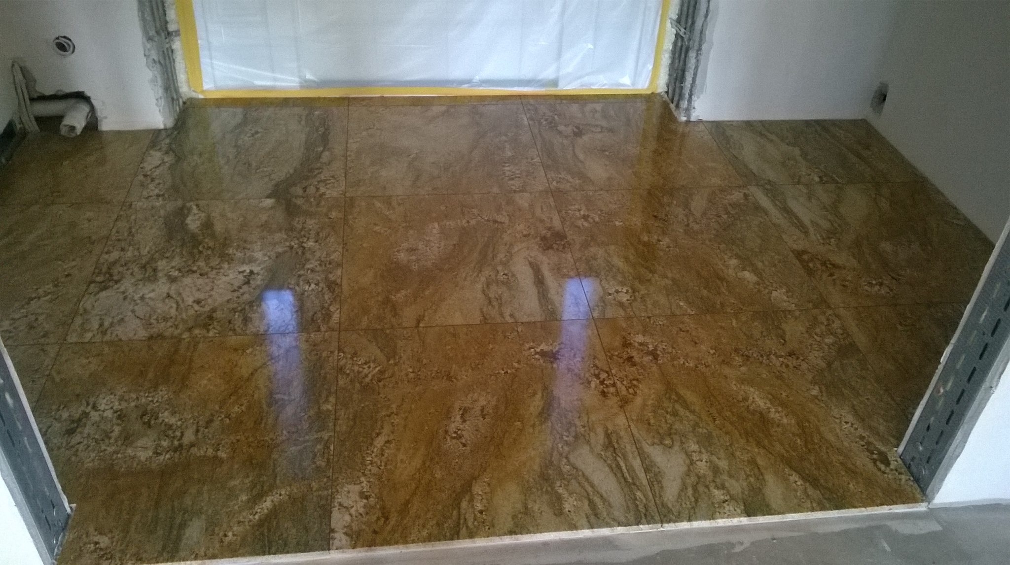 Ułożona, błyszcząca podłoga z płytek imitujących marmur w odcieniach brązu i beżu, widoczne zabezpieczenia folią malarską na ścianie, instalacja hydrauliczna w rogu.