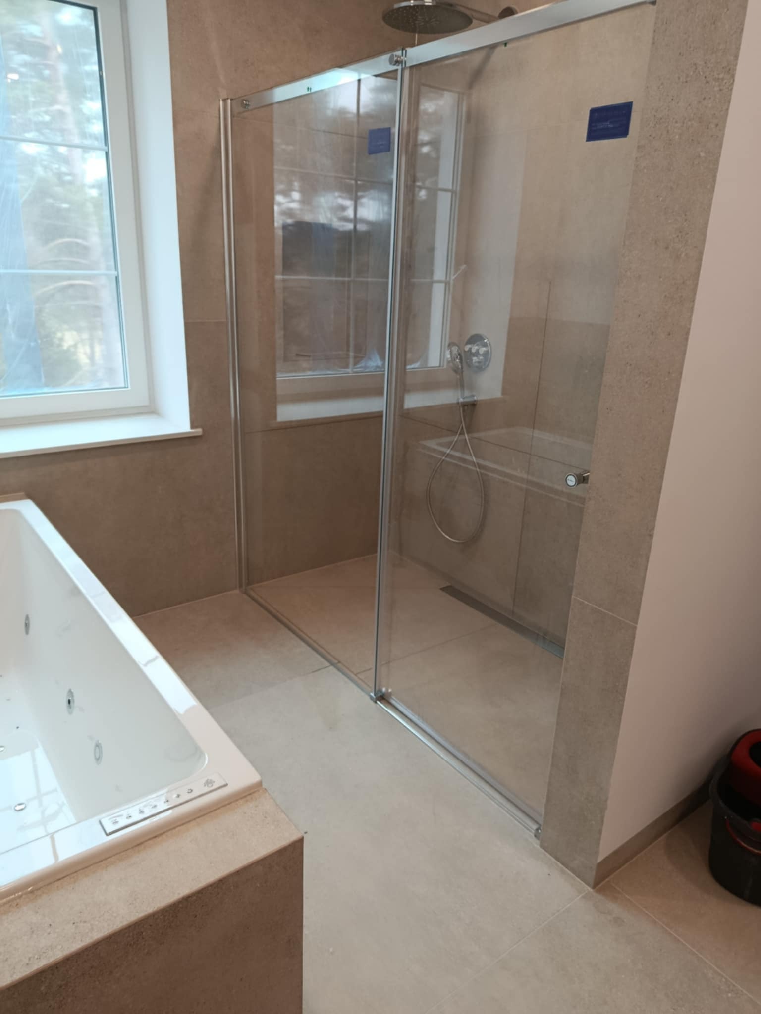 Nowoczesna łazienka z kabiną prysznicową walk-in ze srebrnymi profilami, widok na wannę z hydromasażem i okno z zasłoniętą szybą.