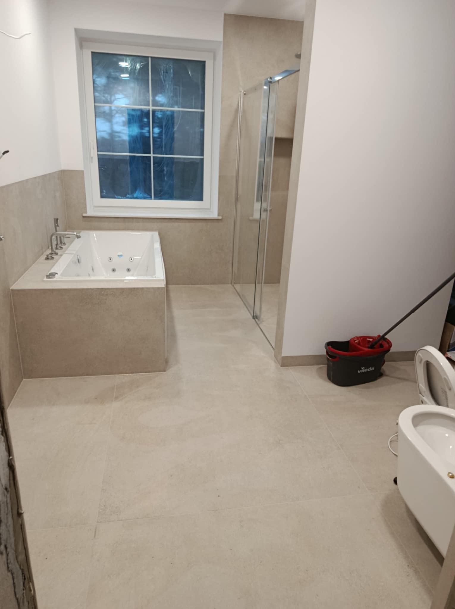 Jasna łazienka po remoncie z wanną z hydromasażem, kabiną prysznicową i bidetem, okno zabezpieczone folią, wiadro i mop w rogu.