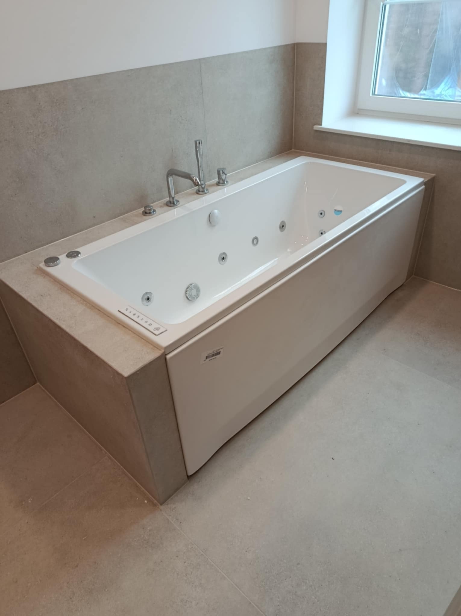 Nowoczesna wanna z hydromasażem w zabudowie z beżowych płytek, chromowana bateria, szare płytki ścienne i podłogowe, okno w tle.