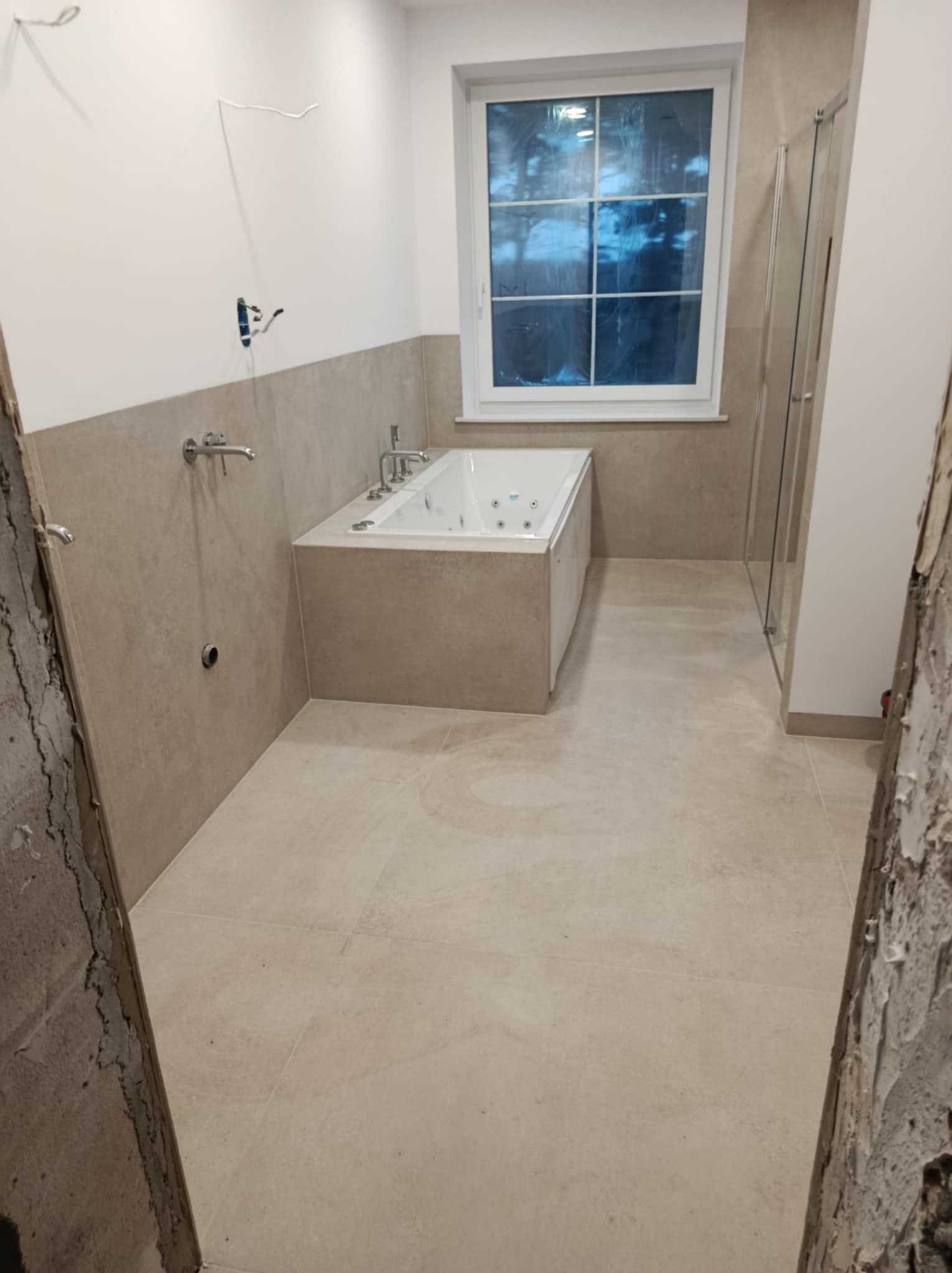 Wnętrze łazienki w trakcie wykańczania w Borne Sulinowo, widoczna wanna z hydromasażem, beżowe płytki na podłodze i ścianach do połowy wysokości, odsłonięte kable i fragment ściany w stanie surowym.