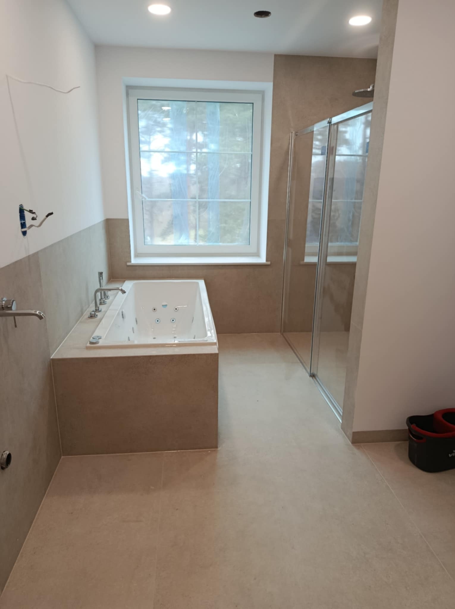 Wanna z hydromasażem, kabina prysznicowa i beżowe płytki w łazience w trakcie wykańczania, widoczne niedokończone podłączenia armatury i wiadro na podłodze.