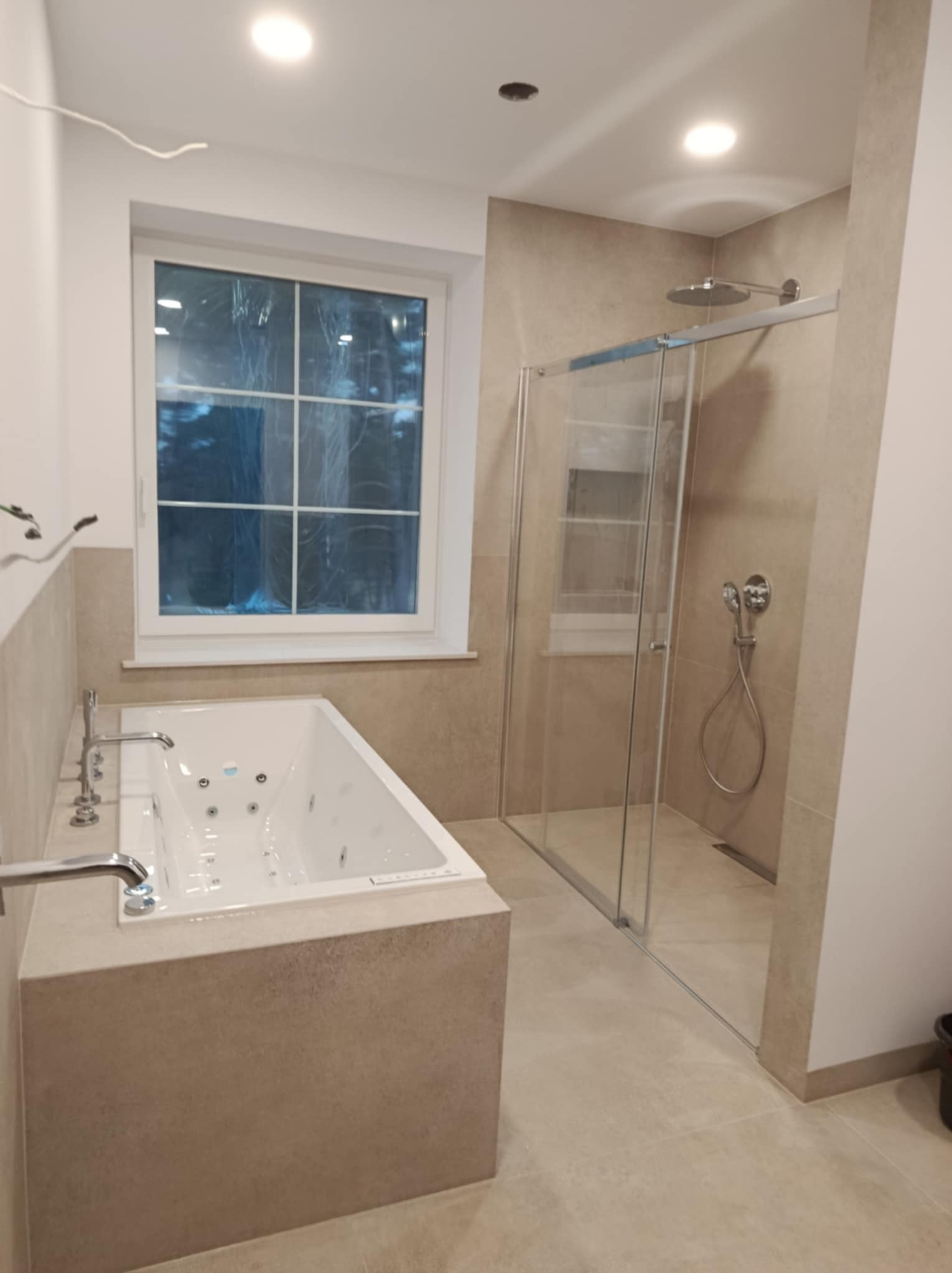 Nowoczesna łazienka z wanną jacuzzi obudowaną płytkami w odcieniach beżu oraz przeszkloną kabiną prysznicową typu walk-in z deszczownicą, widoczne chromowane baterie i okno z folią ochronną.