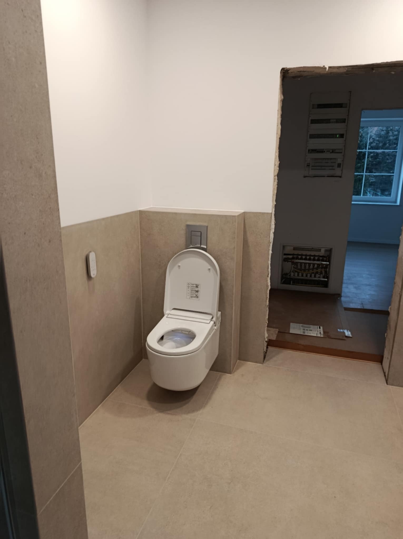 Nowoczesna toaleta myjąca z panelem sterowania na ścianie w wykończonej łazience z beżowymi płytkami, widok na sąsiednie pomieszczenie przez surowy otwór drzwiowy.