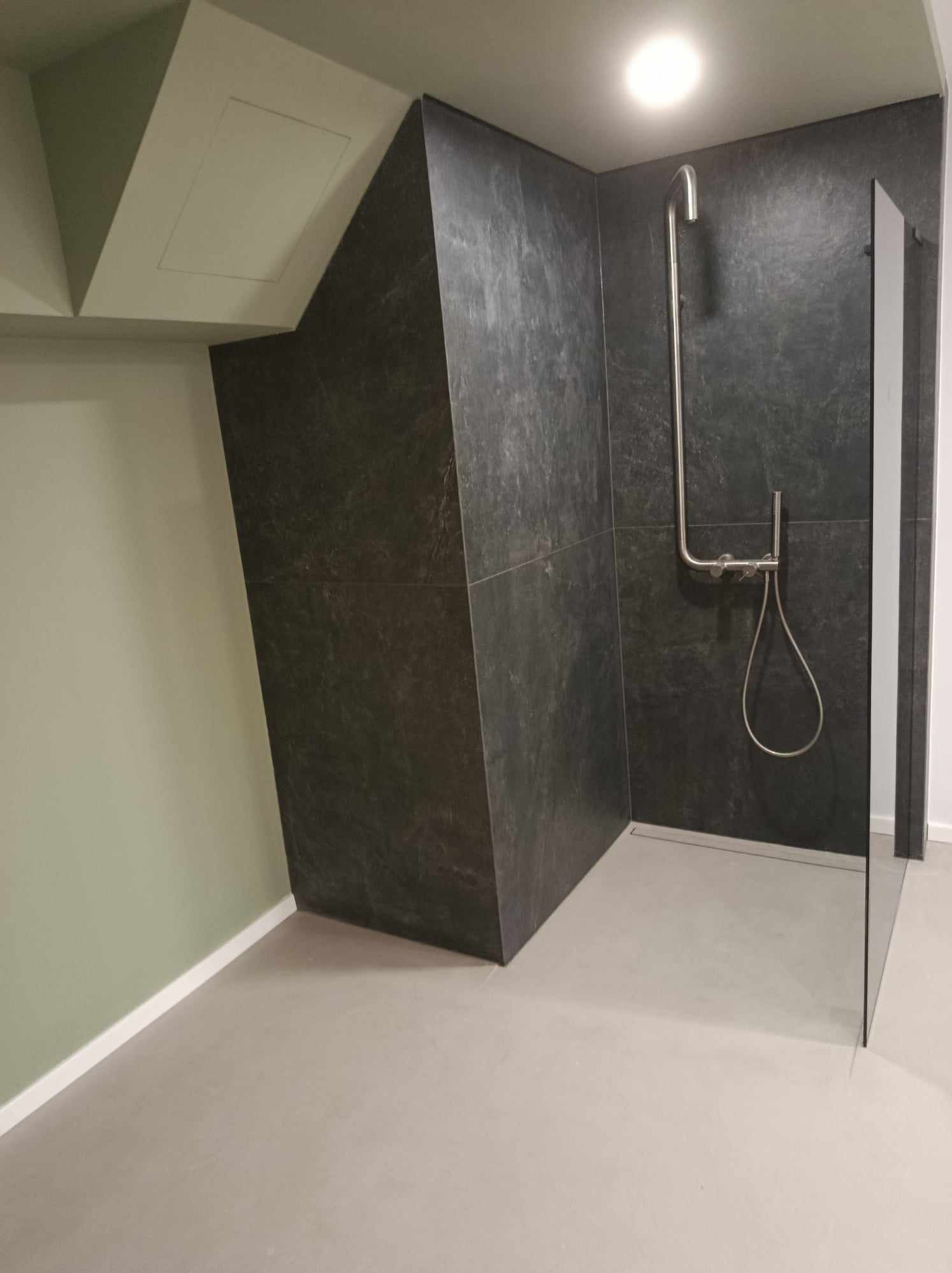 Nowoczesna kabina prysznicowa z ciemnymi płytkami imitującymi kamień, chromowaną armaturą i szklaną ścianką, w łazience ze skośnym sufitem i zielonymi ścianami.