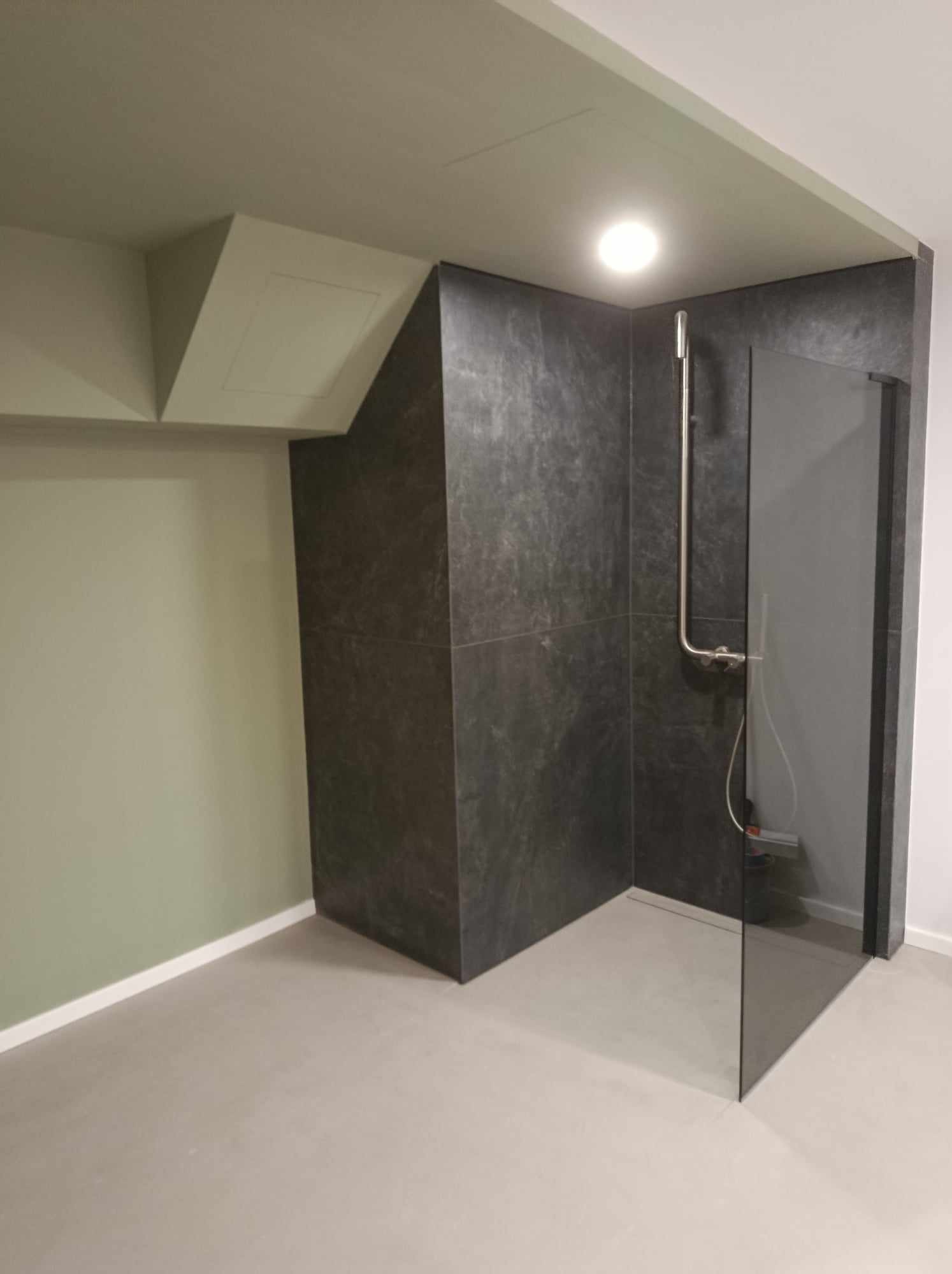 Nowoczesna kabina prysznicowa walk-in z ciemnoszarymi płytkami imitującymi kamień, szklana ścianka bez brodzika, minimalistyczna bateria prysznicowa, oświetlenie sufitowe punktowe, ściana...