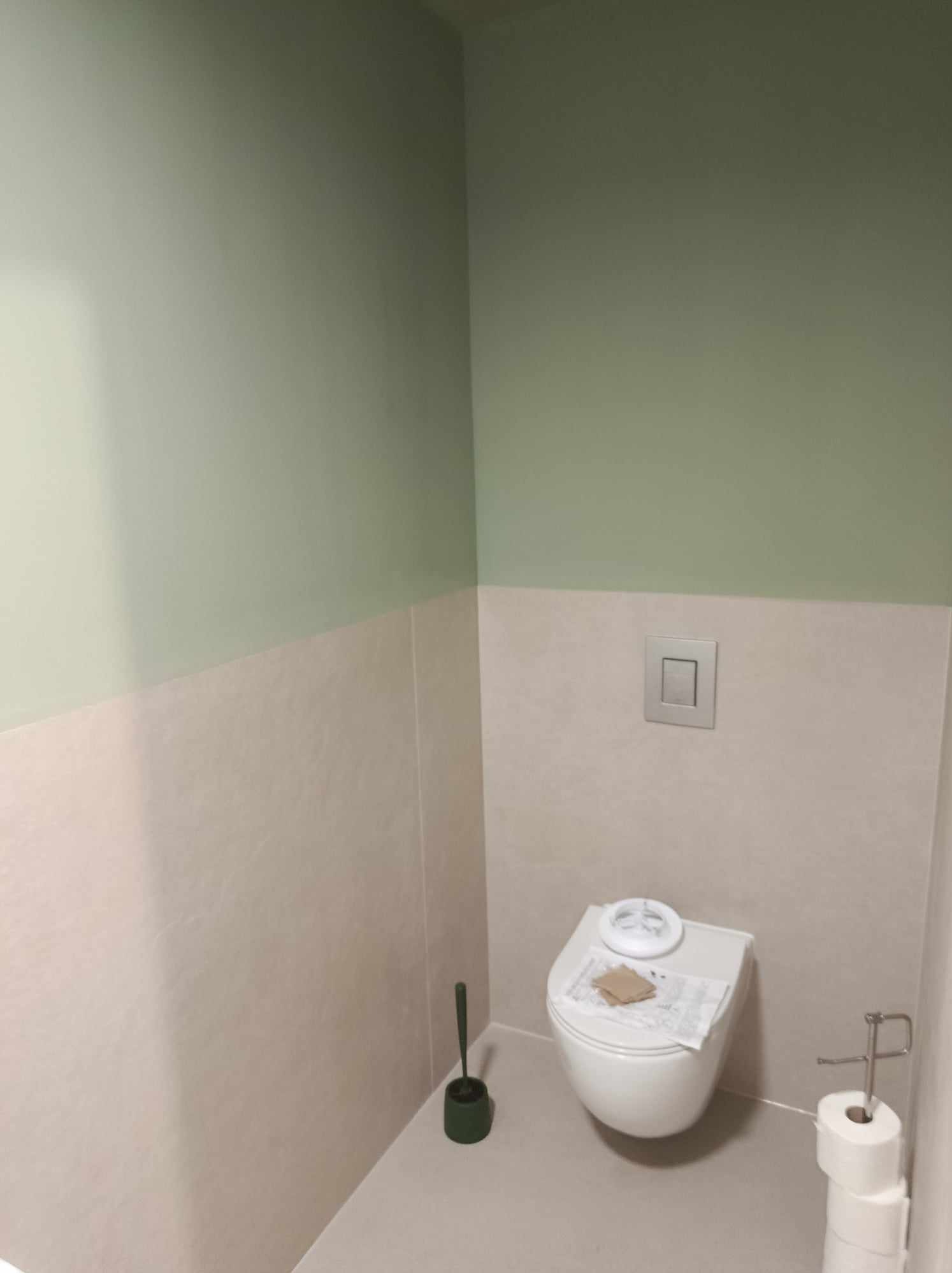 Nowoczesna toaleta podwieszana z deską sedesową z funkcją bidetu, wykończona w dwukolorowej palecie: dolna część ścian w odcieniu beżu, górna w pastelowej zieleni, z minimalistycznym przyciskiem...